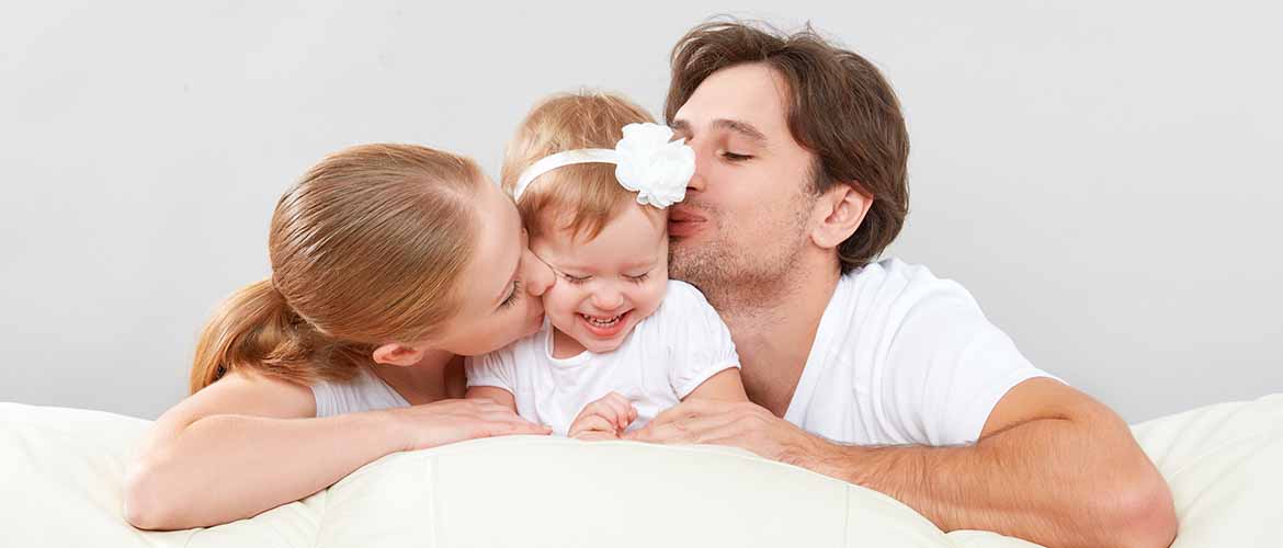 Family of 3, mother, father and baby, all dressed in white, parents kiss the child on the cheek