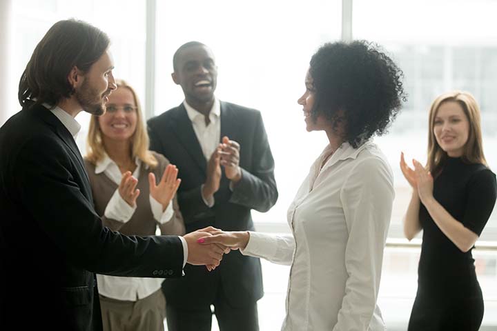 A business man and business woman shaking hands in a conference room with people clapping in the background