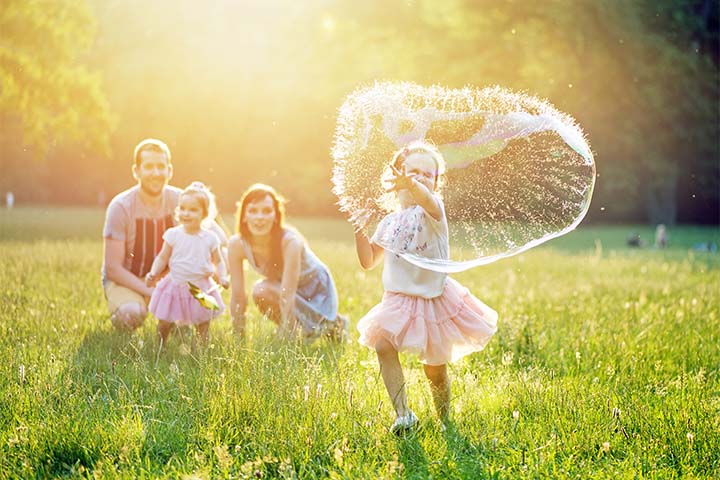 Litle girl playing in a large field blowing bubbles while parents watch and smile