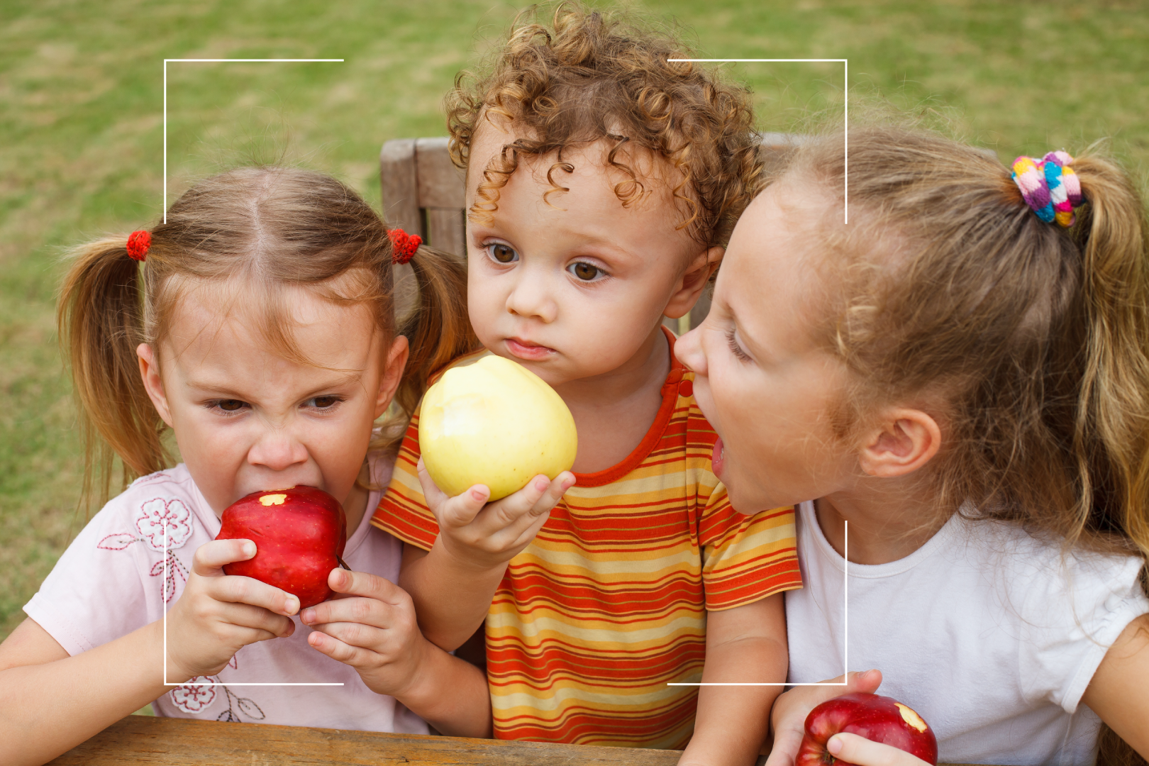 3 niños pequeños comiendo manzanas rojas en una mesa de picnic