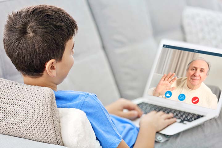 Young boy in a blue shirt laying on a gray couch while video chatting with his grandmother through a tablet